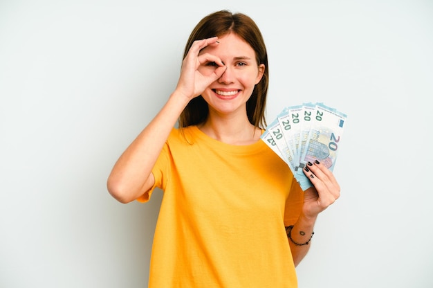 Young English woman holding banknotes isolated on blue background excited keeping ok gesture on eye