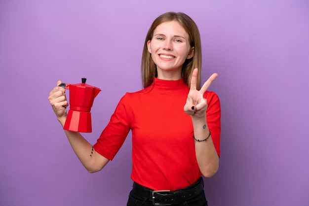 Young English woman holding coffee pot isolated on purple background smiling and showing victory sign