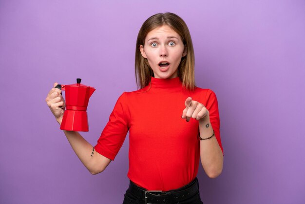 Young English woman holding coffee pot isolated on purple background surprised and pointing front