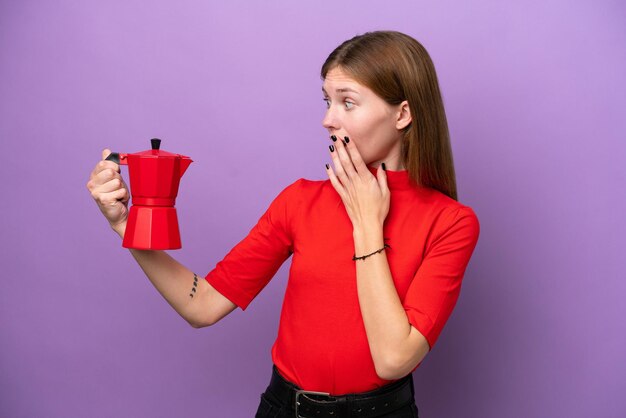 Young English woman holding coffee pot isolated on purple background with surprise and shocked facial expression