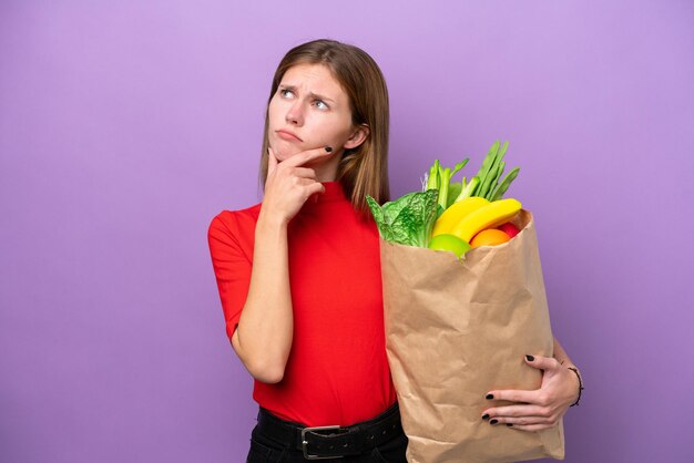 Young english woman holding a grocery shopping bag isolated on purple background having doubts