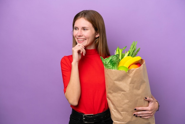 紫色の背景に食料品の買い物袋を持ち、横を見て微笑む若いイギリス人女性