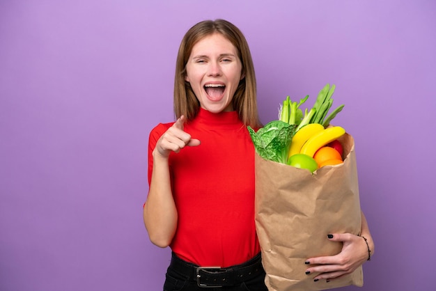 食料品の買い物袋を保持している若い英国人女性は、紫色の背景に驚き、前を向いています