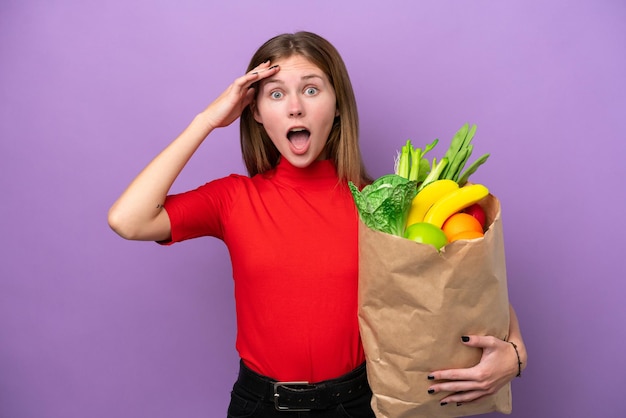 Young english woman holding a grocery shopping bag isolated on purple background with surprise expression