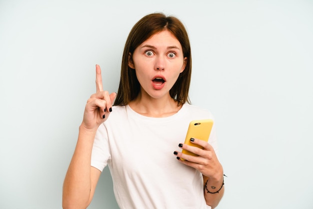 Young English woman holding mobile phone isolated on blue background having an idea, inspiration concept.