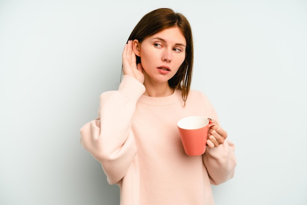 Young english woman holding a mug isolated on blue background\
trying to listening a gossip