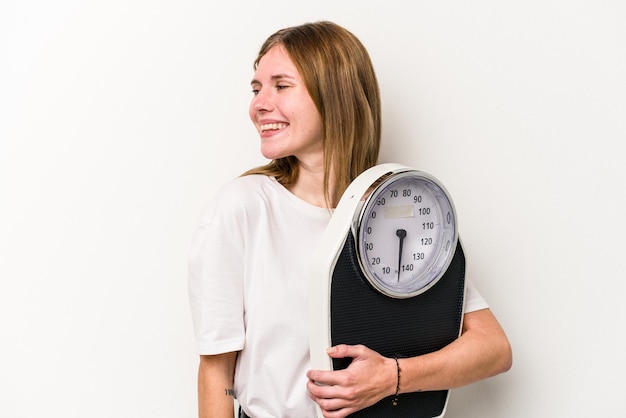 Young English woman holding a scale isolated on white background looks aside smiling cheerful and pleasant