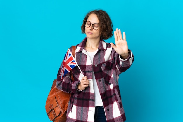 Young English woman holding an United Kingdom flag isolated on blue background making stop gesture and disappointed