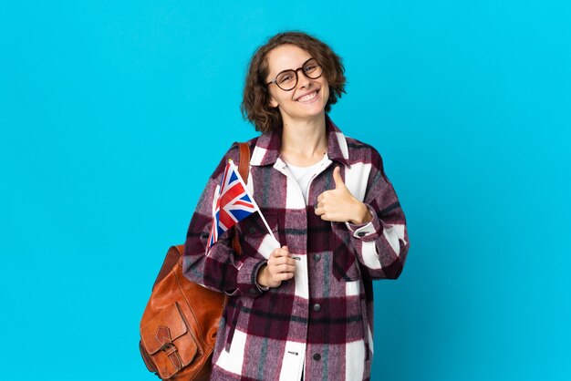 Young English woman holding an United Kingdom flag isolated on blue wall giving a thumbs up gesture