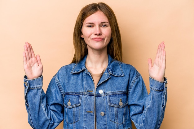 Young English woman isolated on beige background holding something little with forefingers smiling and confident