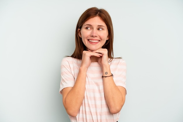 Young English woman isolated on blue background keeps hands under chin, is looking happily aside.