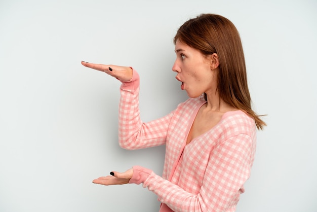 Young English woman isolated on blue background shocked and amazed holding a copy space between hands