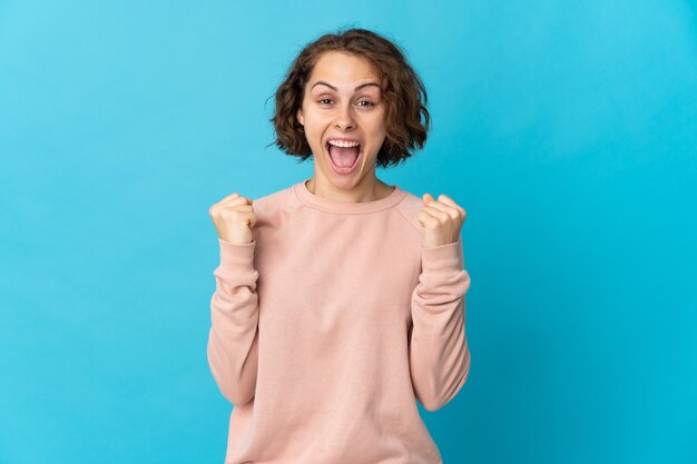 Young English woman isolated on blue wall celebrating a victory in winner position