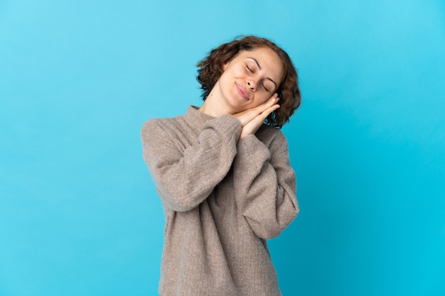 Young English woman isolated on blue wall making sleep gesture in dorable expression