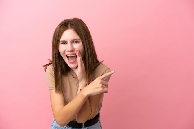 Young English woman isolated on pink background pointing to the side to present a product and whispering something