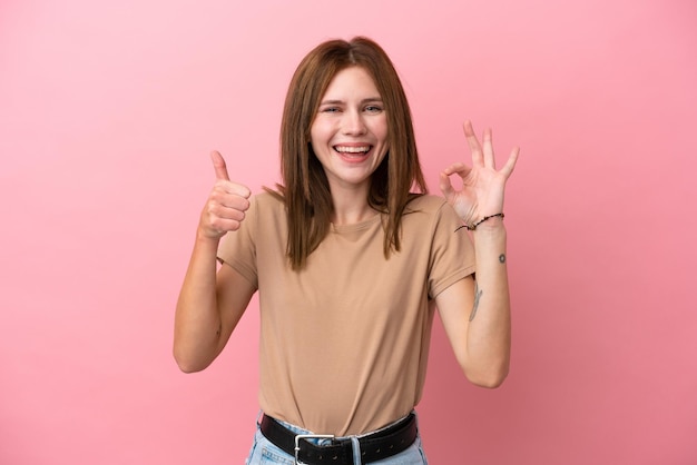 Молодая англичанка, изолированная на розовом фоне, показывает знак "ОК" и жест "большой палец вверх"