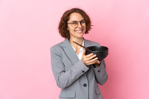 Young English woman isolated on pink wall holding a bowl of noodles with chopsticks