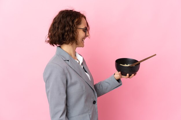 Young English woman isolated on pink wall with happy expression while holding a bowl of noodles with chopsticks