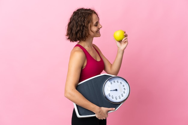 Young English woman isolated on pink wall with weighing machine and with an apple