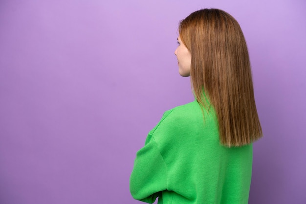 Young english woman isolated on purple background in back\
position and looking back