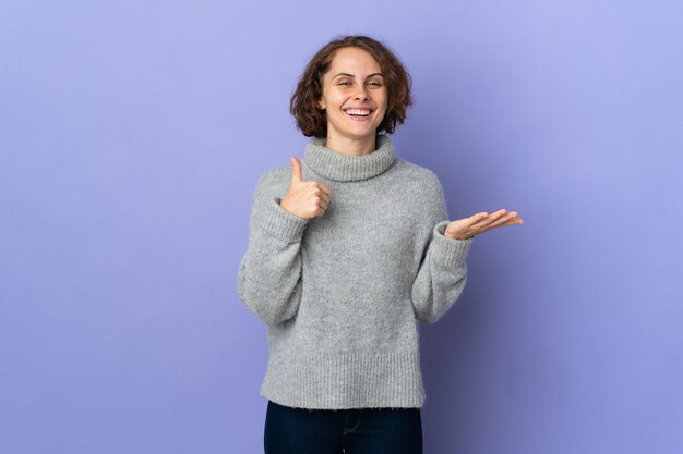 Young English woman isolated on purple background holding copyspace imaginary on the palm to insert an ad and with thumbs up