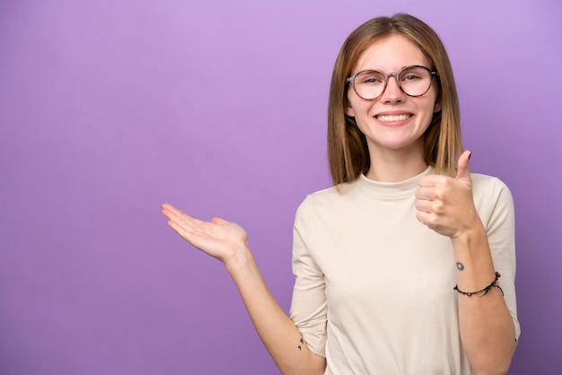 Young English woman isolated on purple background holding copyspace imaginary on the palm to insert an ad and with thumbs up