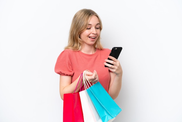 Young English woman isolated on white background holding shopping bags and writing a message with her cell phone to a friend