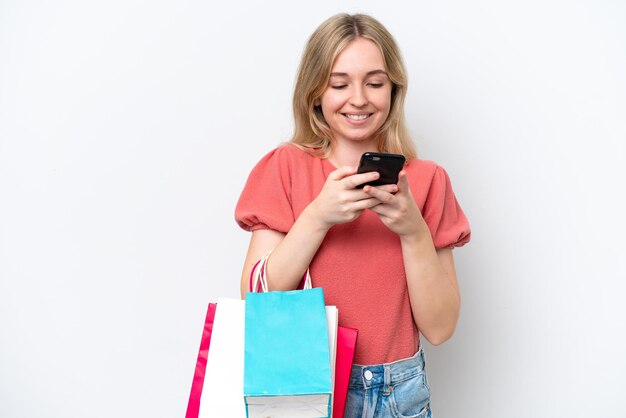 Young English woman isolated on white background holding shopping bags and writing a message with her cell phone to a friend