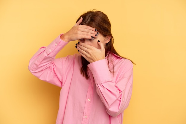 Photo young english woman isolated on yellow background blink at the camera through fingers, embarrassed covering face.