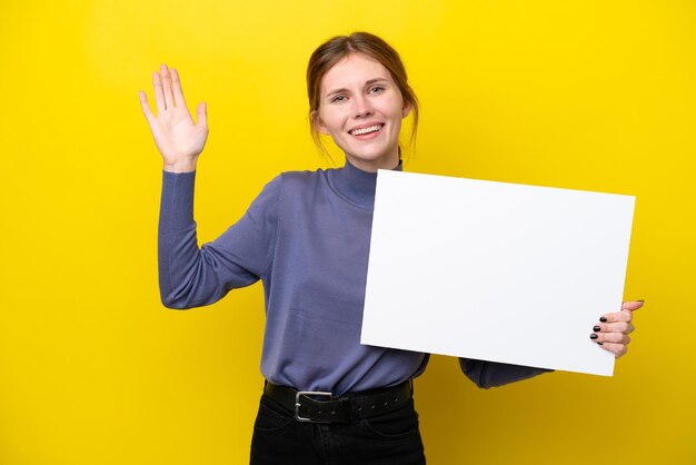 Young English woman isolated on yellow background holding an empty placard and saluting