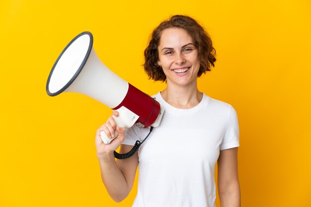 Young English woman isolated on yellow background holding a megaphone and smiling a lot