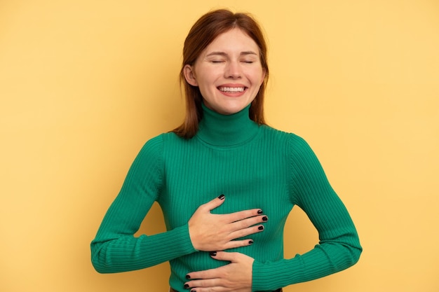 Young English woman isolated on yellow background laughs happily and has fun keeping hands on stomach.