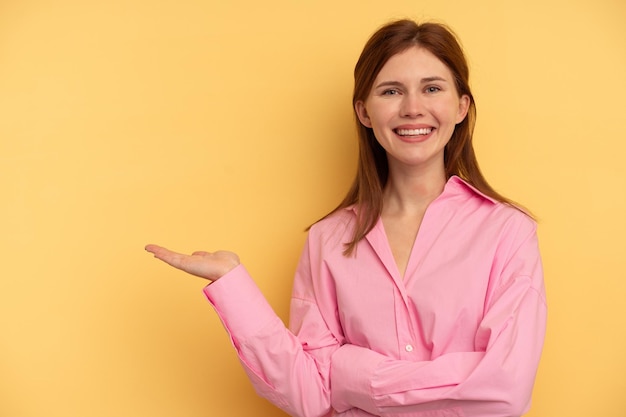 Young English woman isolated on yellow background showing a copy space on a palm and holding another hand on waist.