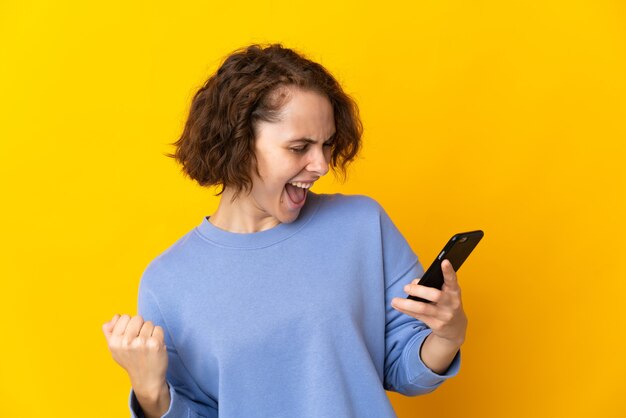 Young English woman isolated on yellow background using mobile phone and doing victory gesture