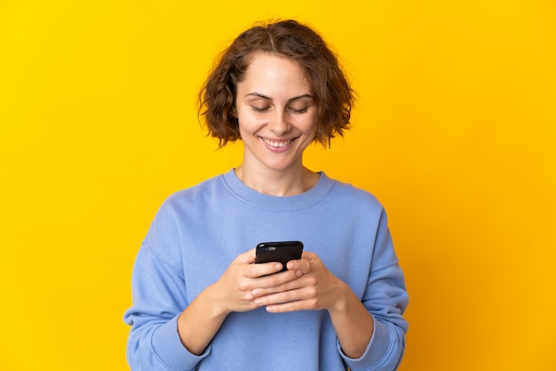 Молодая англичанка изолирована на желтом пространстве, отправляя сообщение или электронное письмо с мобильного телефона