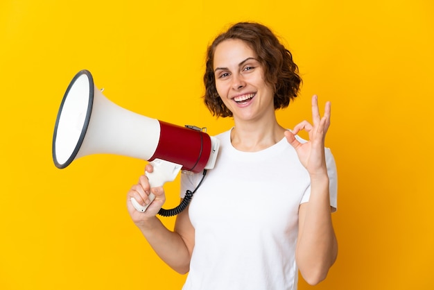 Young English woman isolated on yellow wall holding a megaphone and showing ok sign with fingers