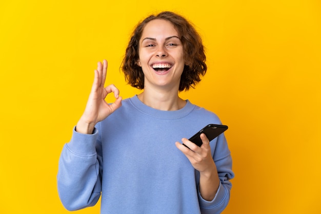 Young english woman isolated on yellow wall using mobile phone and doing ok sign