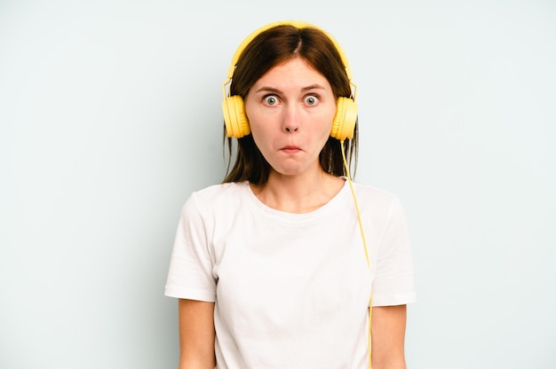 Young English woman listening to music isolated on blue background shrugs shoulders and open eyes confused