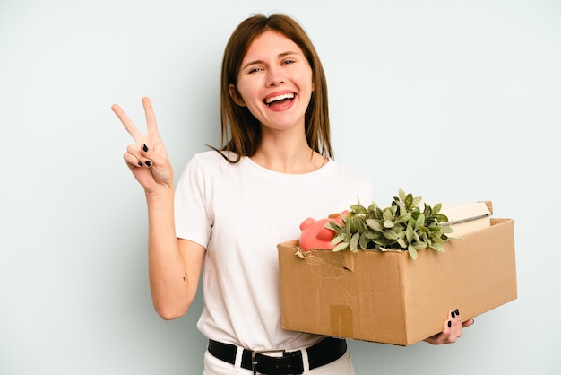 Young English woman making a move while picking up a box full of things isolated on blue background joyful and carefree showing a peace symbol with fingers.