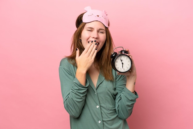 Young English woman in pajamas isolated on pink background in pajamas and holding clock with surprised expression