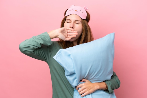 Young English woman in pajamas isolated on pink background in pajamas and holding a pillow and yawning