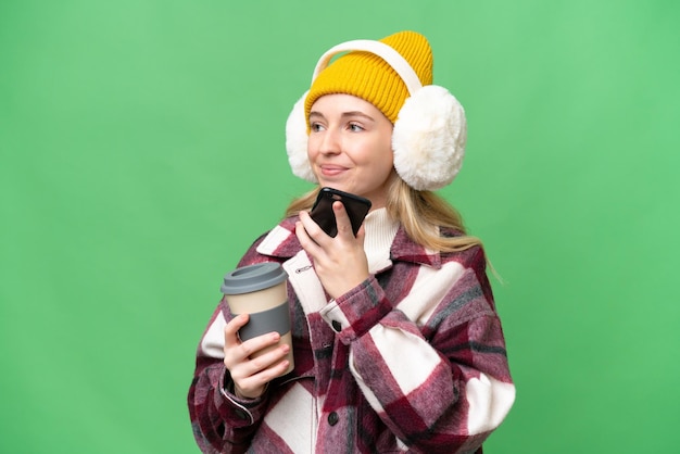 외진 배경 위에 겨울 머프를 입은 젊은 영국 여성이 커피를 들고 모바일을 들고 있습니다.