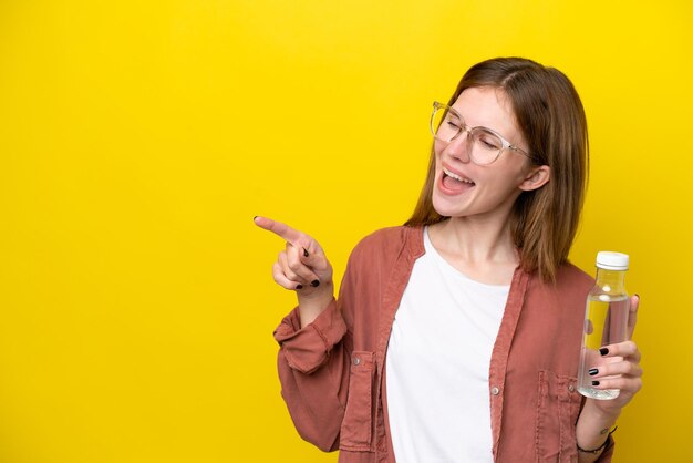 Young English woman with a bottle of water isolated on yellow background pointing finger to the side and presenting a product