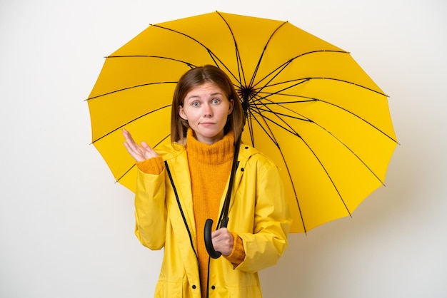 Молодая англичанка в непромокаемом пальто и зонтике на белом фоне сомневается, поднимая руки