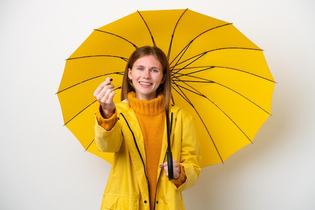 お金のジェスチャーを作る白い背景で隔離の防雨コートと傘を持つ若いイギリス人女性