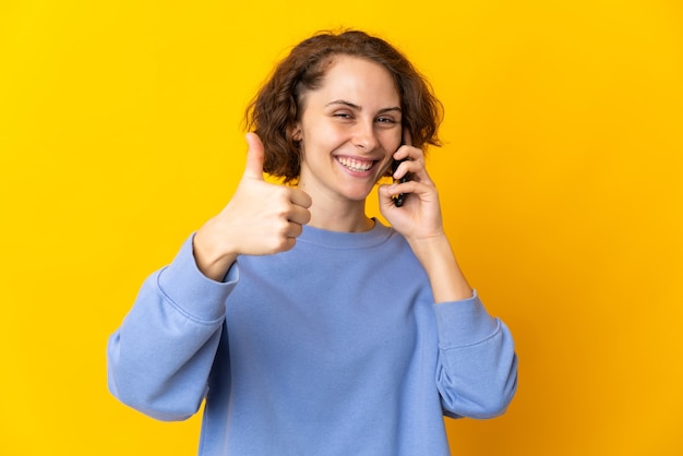 Young English woman on yellow keeping a conversation with the mobile while doing thumbs up