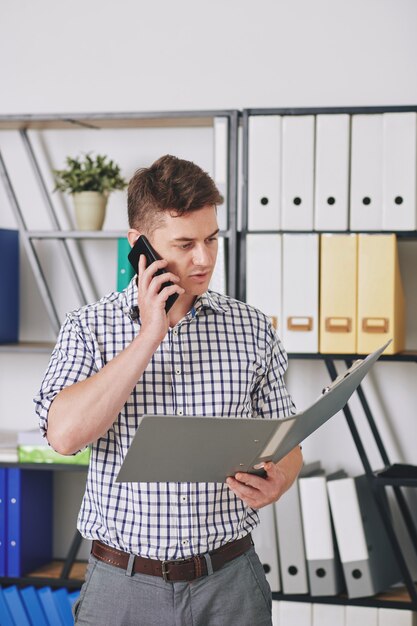 Young entrepreneur reading document in opened folder and talking on phone with client or coworker