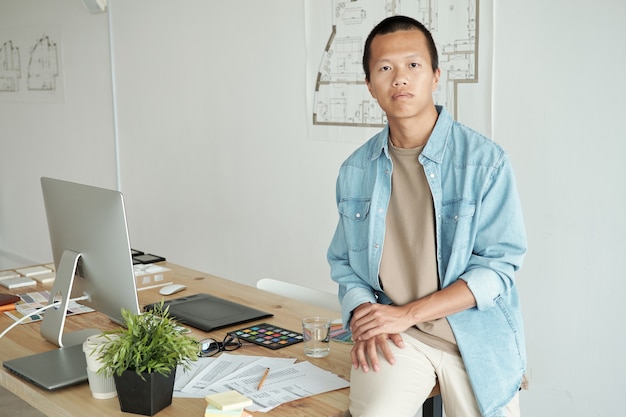 Молодой энергичный китайский бизнесмен сидит за столом с бумагами, цветовой палитрой, образцами, графическим планшетом и монитором компьютера