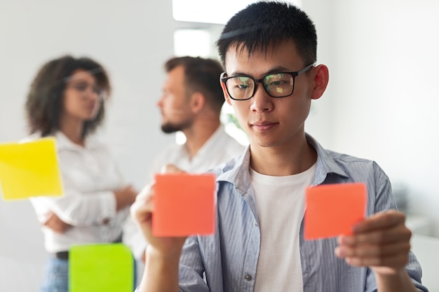 Young ethnic male in casual outfit and eyeglasses putting paper sticky notes on glass wall while having meeting with startup colleagues and discussing business strategy