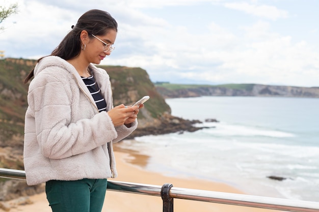 砂浜の近くでスマートフォンでチャットする若い民族の女性